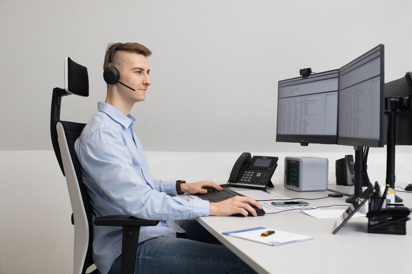 Junger Mann im hellblauen Hemd mit Headset sitzt an einem aufgeräumten Schreibtisch und arbeitet konzentriert am Computer. Vor ihm stehen zwei Monitore mit Datentabellen, daneben ein Tischtelefon. Er bedient Maus und Tastatur – typische Szene aus dem Kund