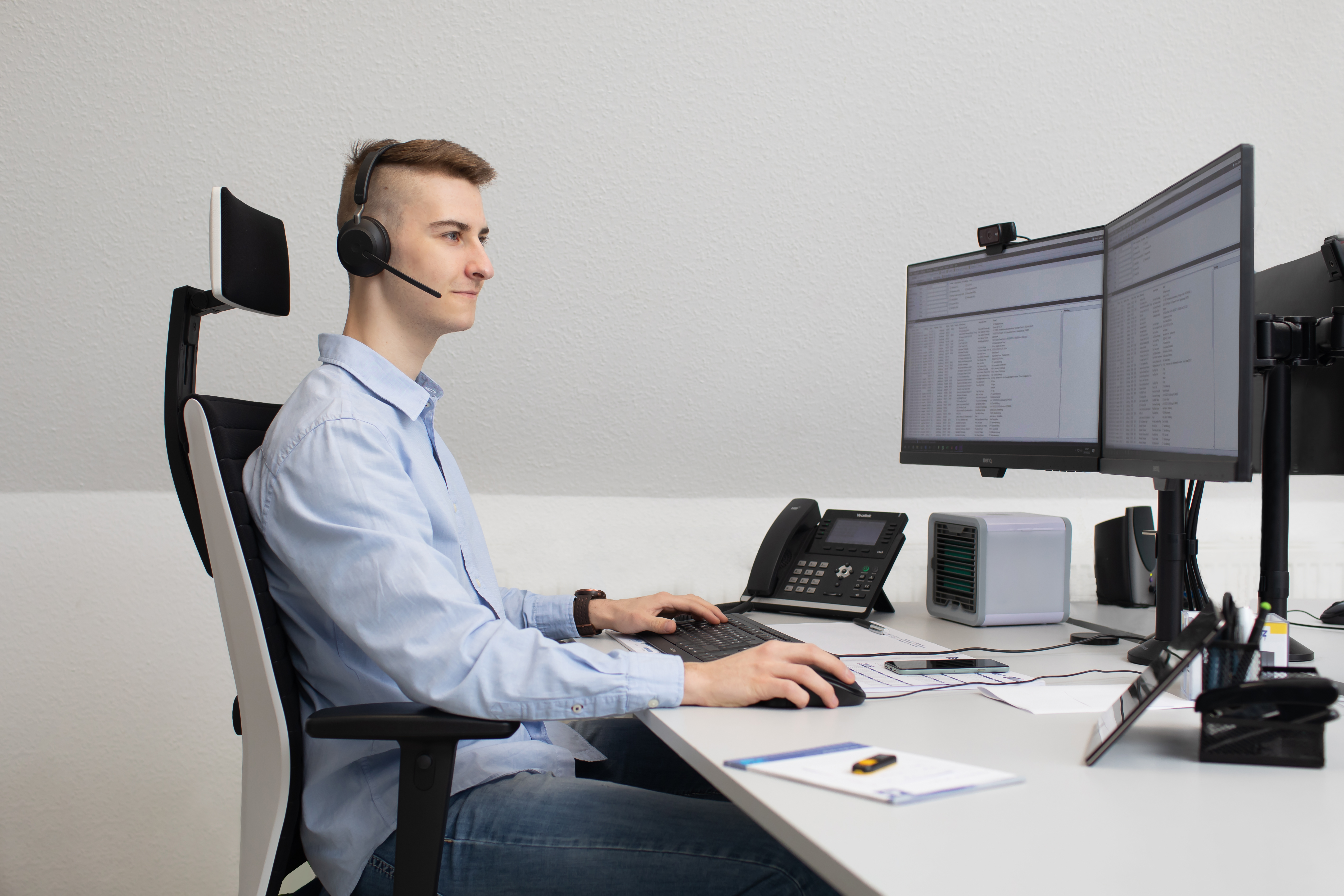 Junger Mann im hellblauen Hemd mit Headset sitzt an einem aufgeräumten Schreibtisch und arbeitet konzentriert am Computer. Vor ihm stehen zwei Monitore mit Datentabellen, daneben ein Tischtelefon. Er bedient Maus und Tastatur – typische Szene aus dem Kund